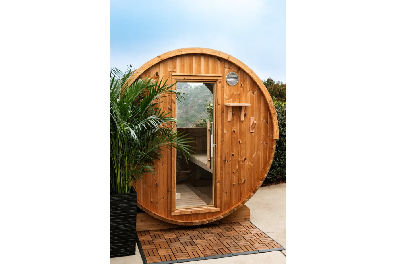 Cylindrical wooden barrel sauna with rounded roof, large glass window, open door, and ventilation vent, positioned on wooden 