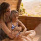 Child wearing blue and yellow floral halter swimsuit sitting in wooden sauna with adult, viewed from inside barrel sauna inte