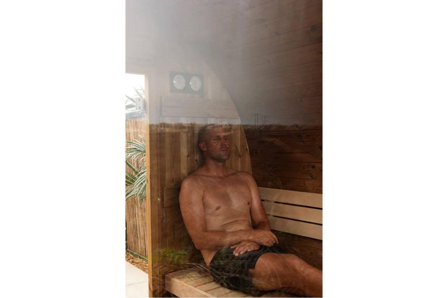 Man relaxing shirtless inside wooden barrel sauna with curved benches and glass door