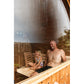 Man and child sitting inside wooden barrel sauna with steaming glass door, viewed from exterior