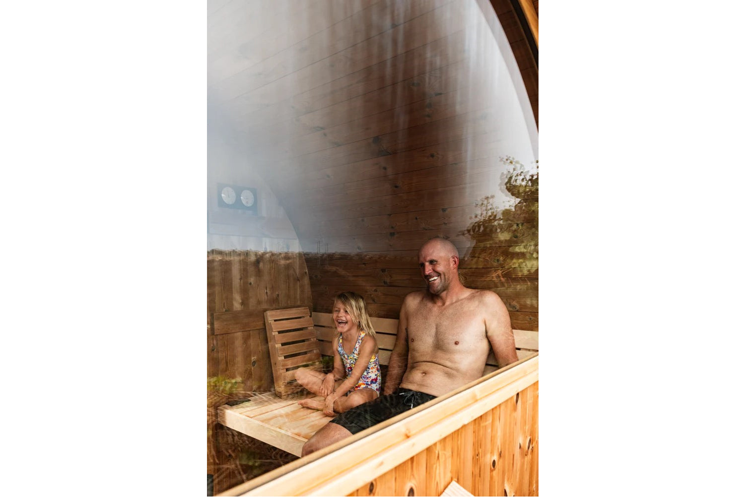 Man and child sitting inside wooden barrel sauna with steaming glass door, viewed from exterior