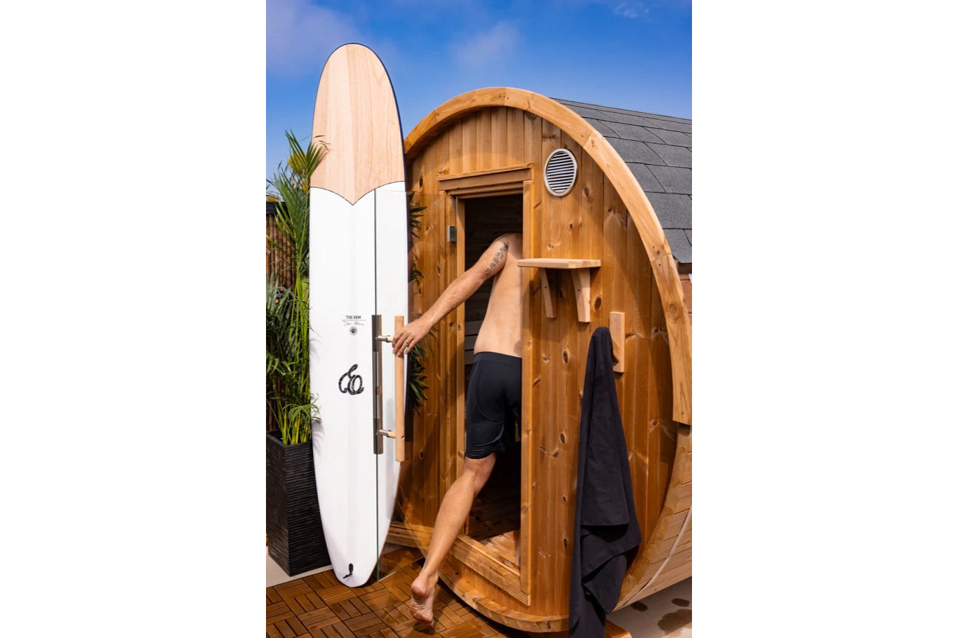 White and wood surfboard leaning against wooden barrel sauna, man in black shorts entering sauna door, blue sky background