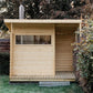 Natural pine wood sauna with large window, open right side, and metal chimney, positioned in backyard garden setting