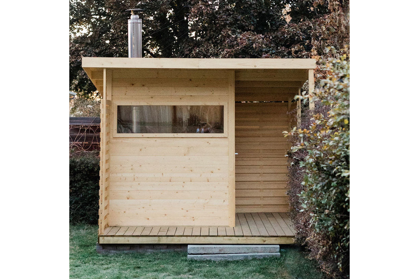 Natural pine wood sauna with large window, open right side, and metal chimney, positioned in backyard garden setting