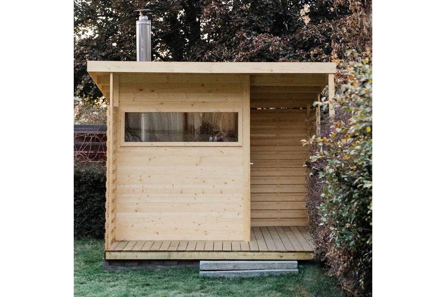 Natural pine wood sauna with large window, open right side, and metal chimney, positioned in backyard garden setting
