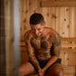 Man with full-body tattoos sitting shirtless in wooden sauna with light wood paneling