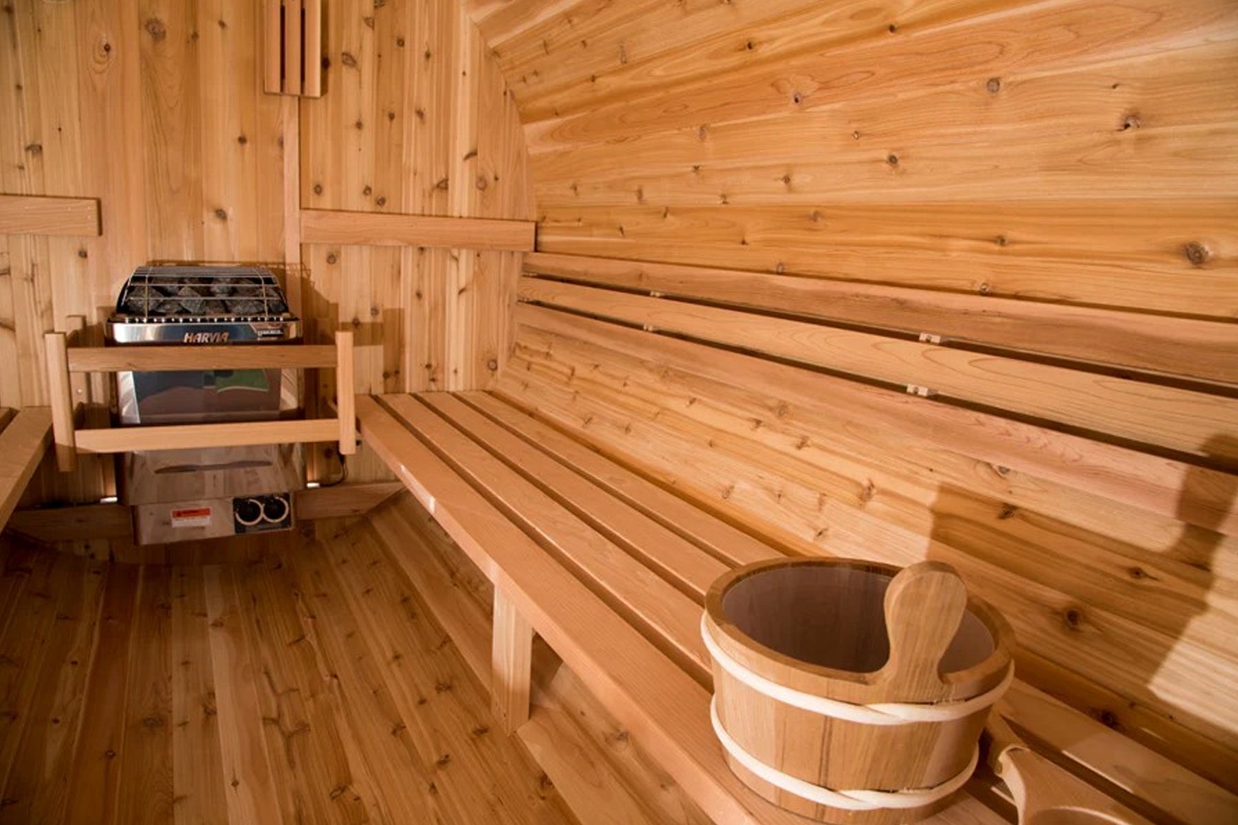 Interior of wooden sauna with benches, cedar walls, barrel heater, and wooden bucket on bench