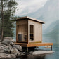 Modern wooden sauna with glass doors and windows on wooden deck, positioned at lakeside with mountains in background