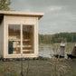 Natural wood outdoor sauna with glass double doors and interior benches, positioned by a lake with two deck chairs nearby
