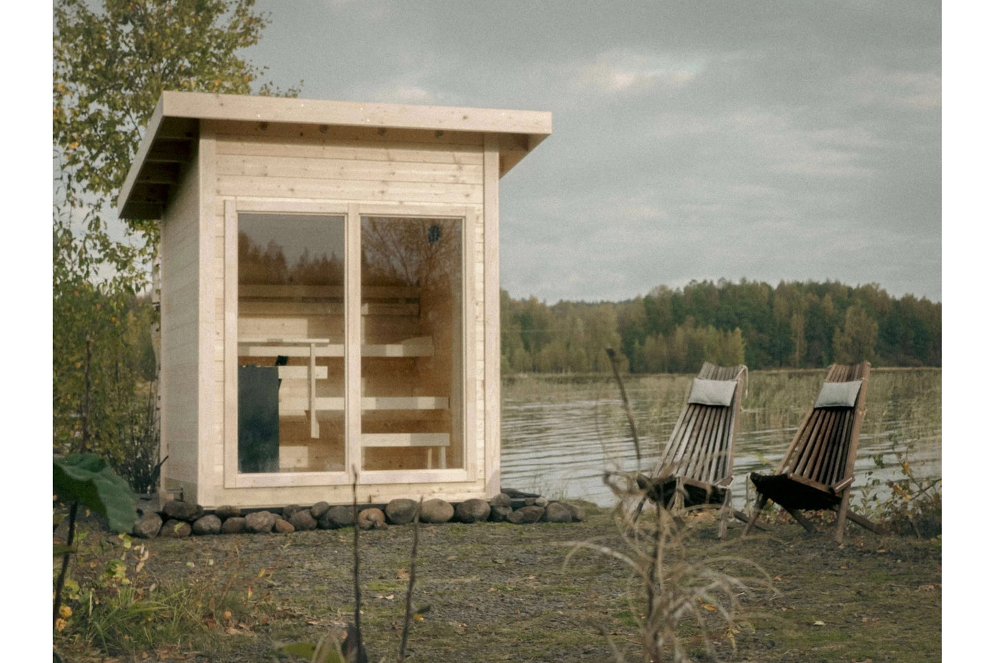 Natural wood outdoor sauna with glass double doors and interior benches, positioned by a lake with two deck chairs nearby