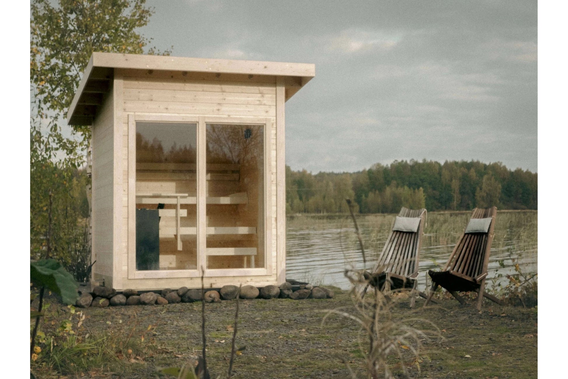 Natural wood outdoor sauna with glass double doors and interior benches, positioned by a lake with two deck chairs nearby