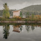 Blush pink wooden sauna with flat roof on lake shore, surrounded by evergreen forest, reflected in still water