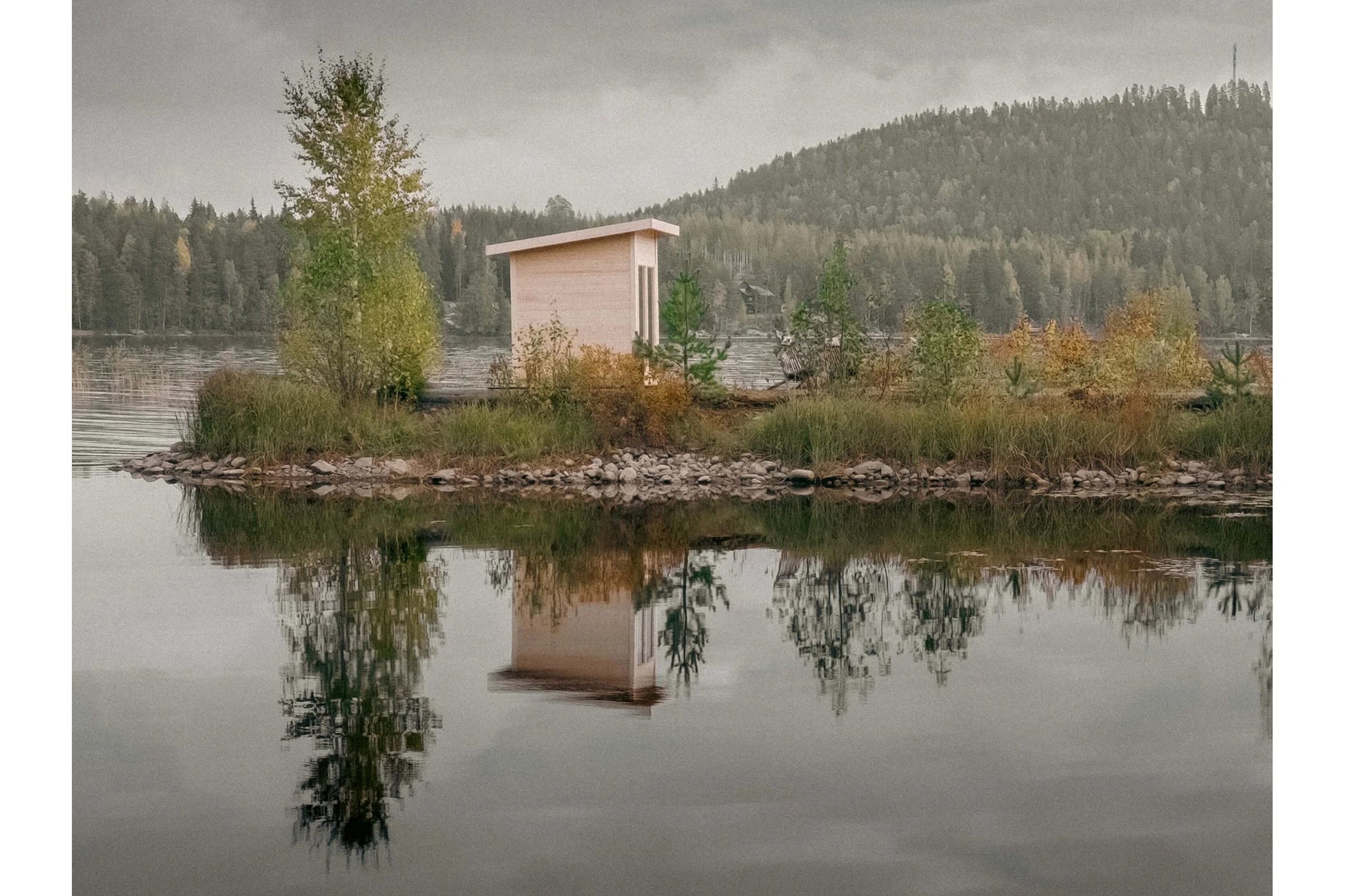 Blush pink wooden sauna with flat roof on lake shore, surrounded by evergreen forest, reflected in still water