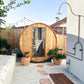 Almost Heaven Watoga barrel sauna with curved wooden exterior and glass door in backyard setting with potted plants