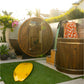 Dark wood barrel sauna with glass door and metal bands, displayed in outdoor garden setting with yellow surfboard and orange 