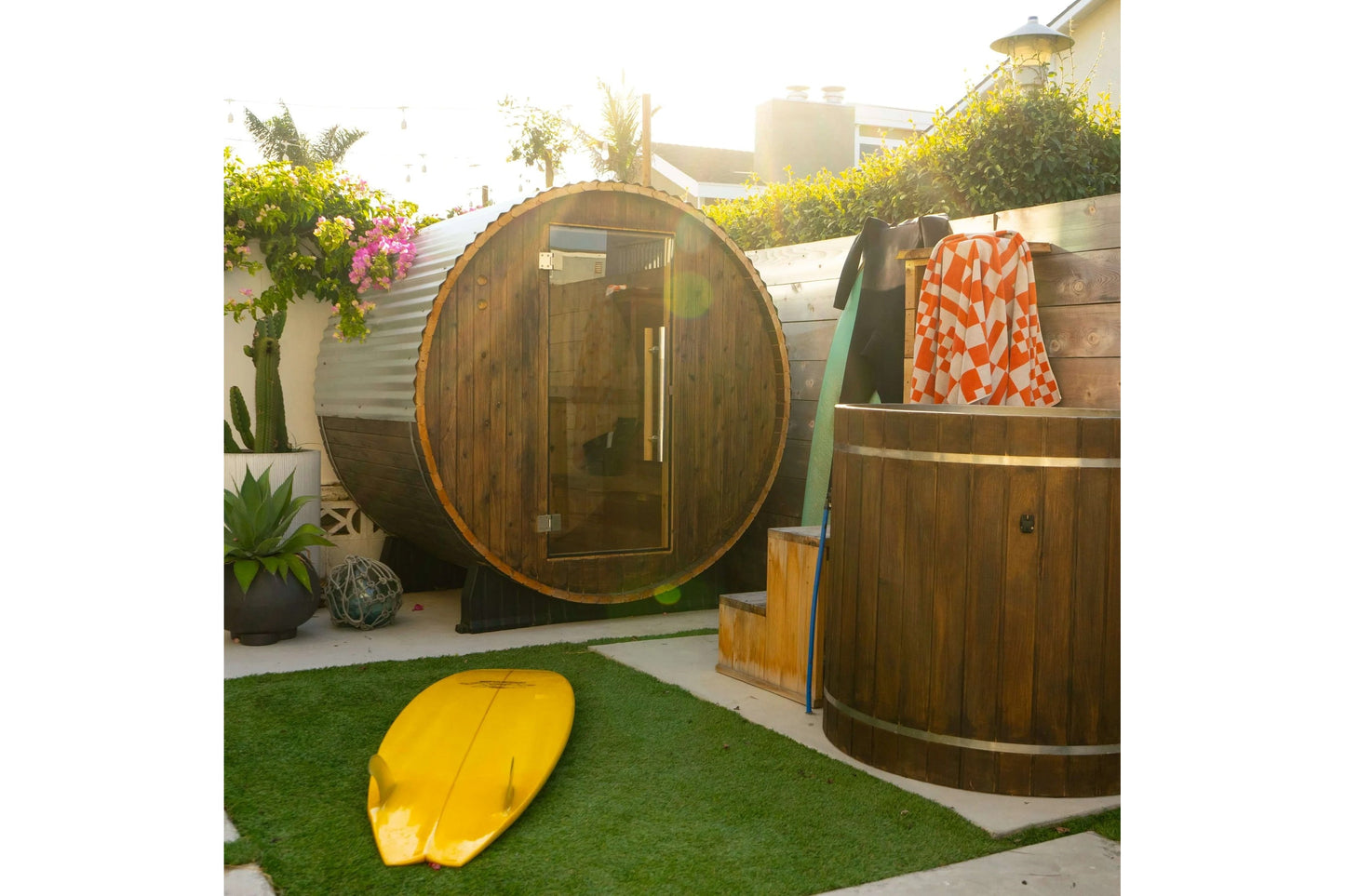 Dark wood barrel sauna with glass door and metal bands, displayed in outdoor garden setting with yellow surfboard and orange 