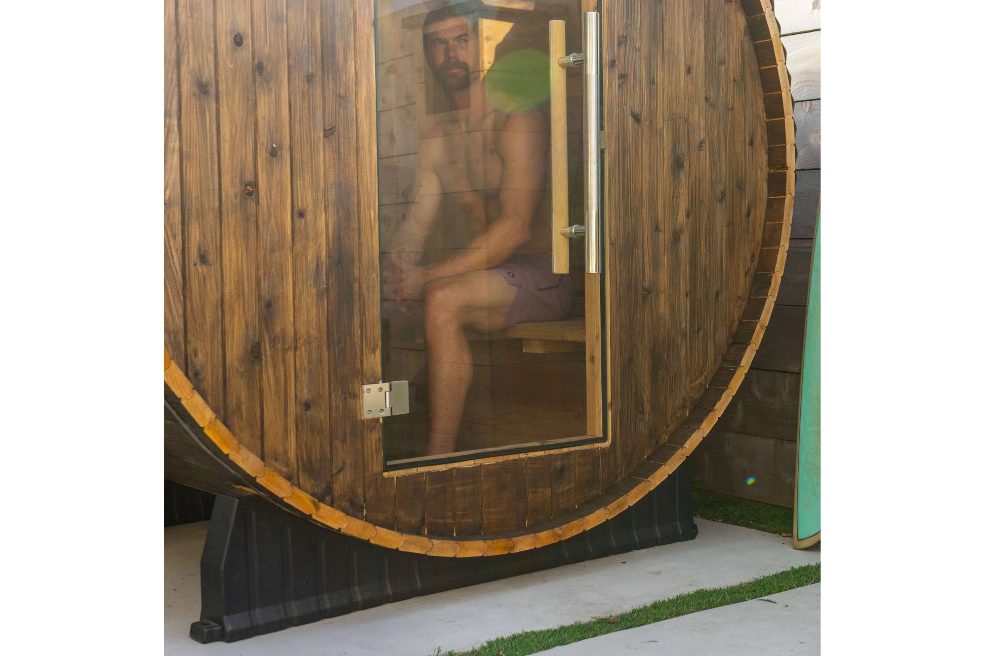 Wooden barrel sauna with glass door, man sitting inside wearing green shorts, natural wood construction with metal bands and 