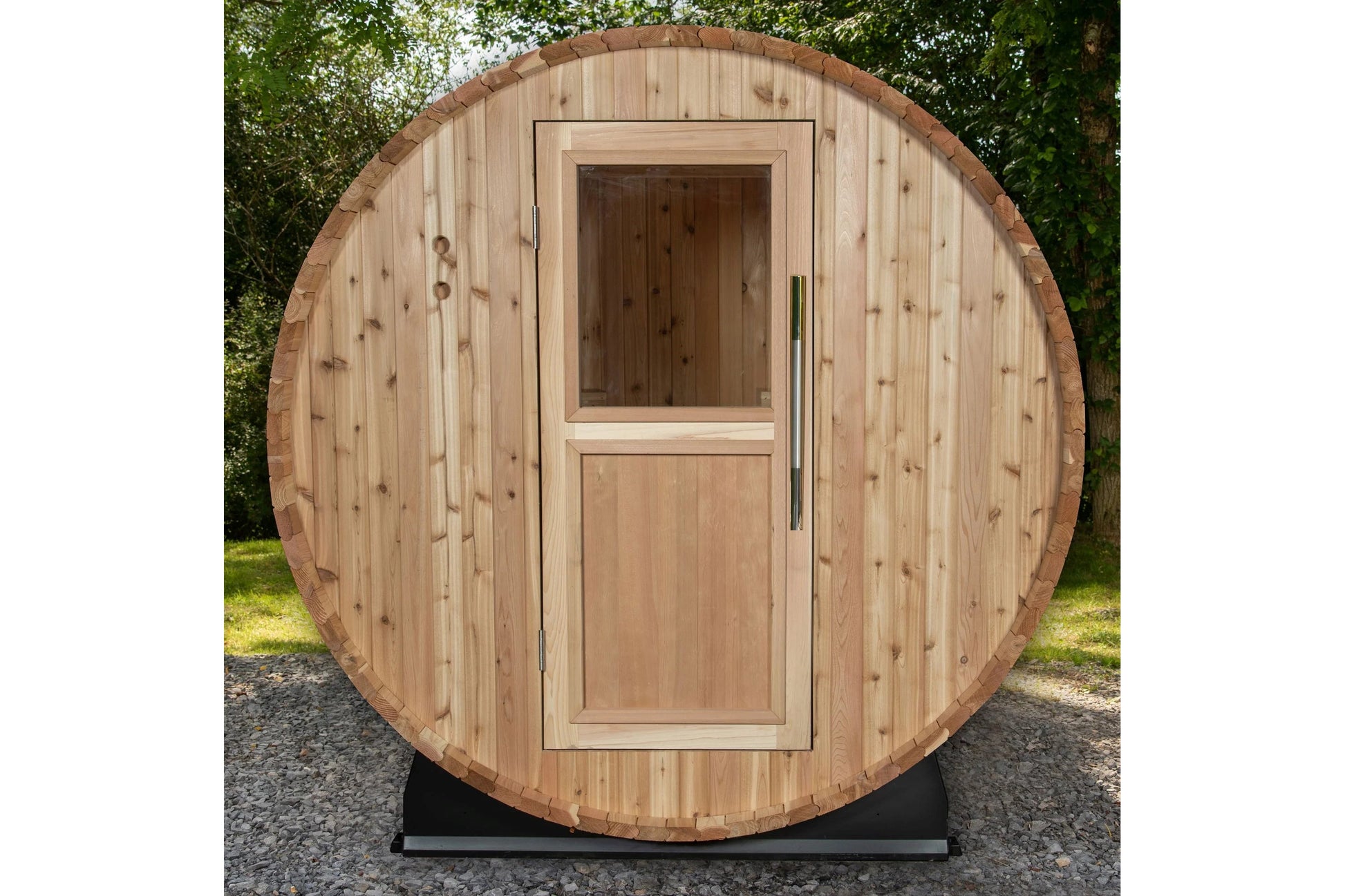 Natural wood barrel sauna with rounded cedar exterior, glass window, and wooden door with handle, displayed outdoors on grave
