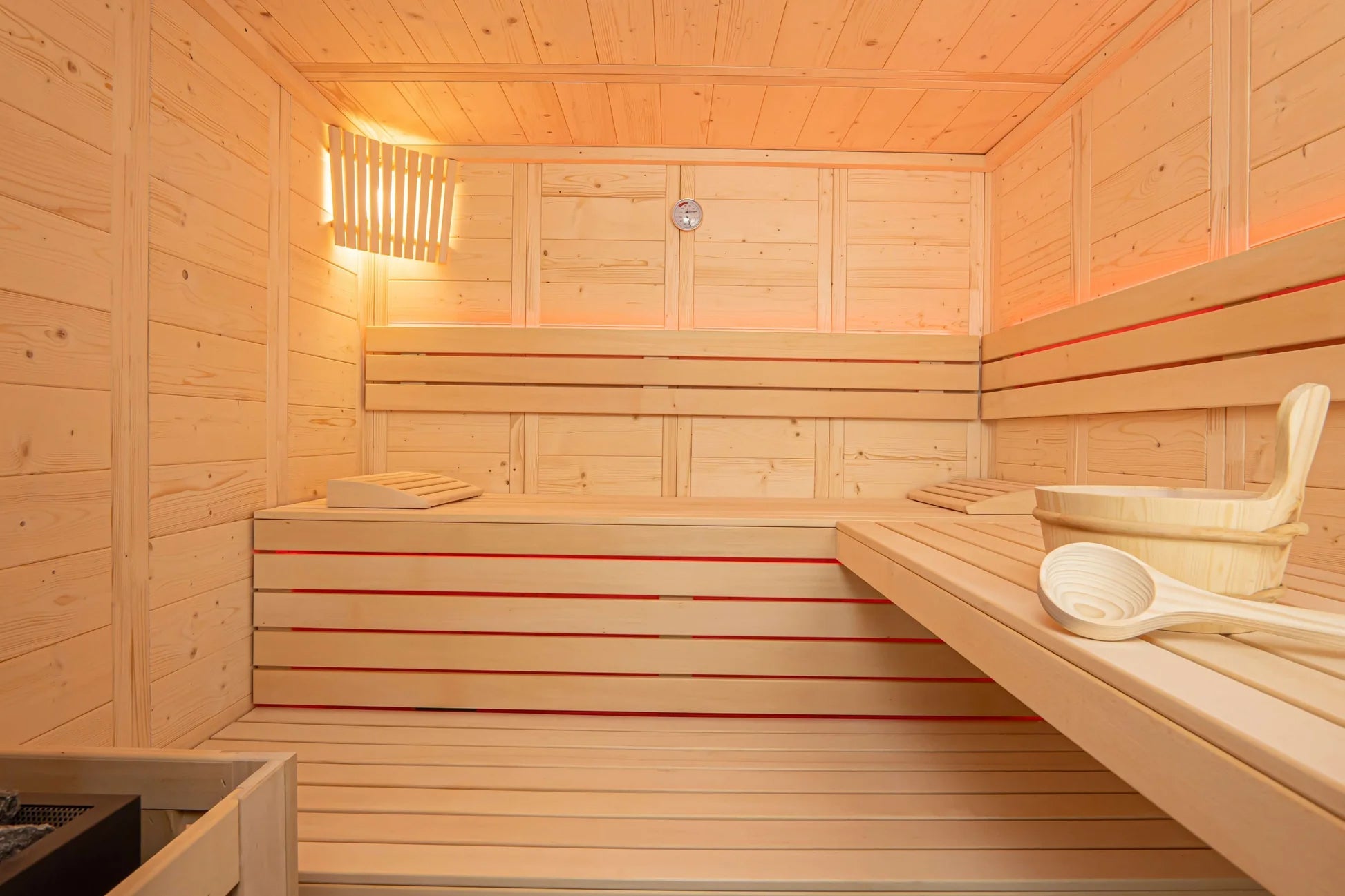 Interior view of light wood sauna with tiered benches, red accent stripe, wooden bucket, ladle, and window with warm lighting