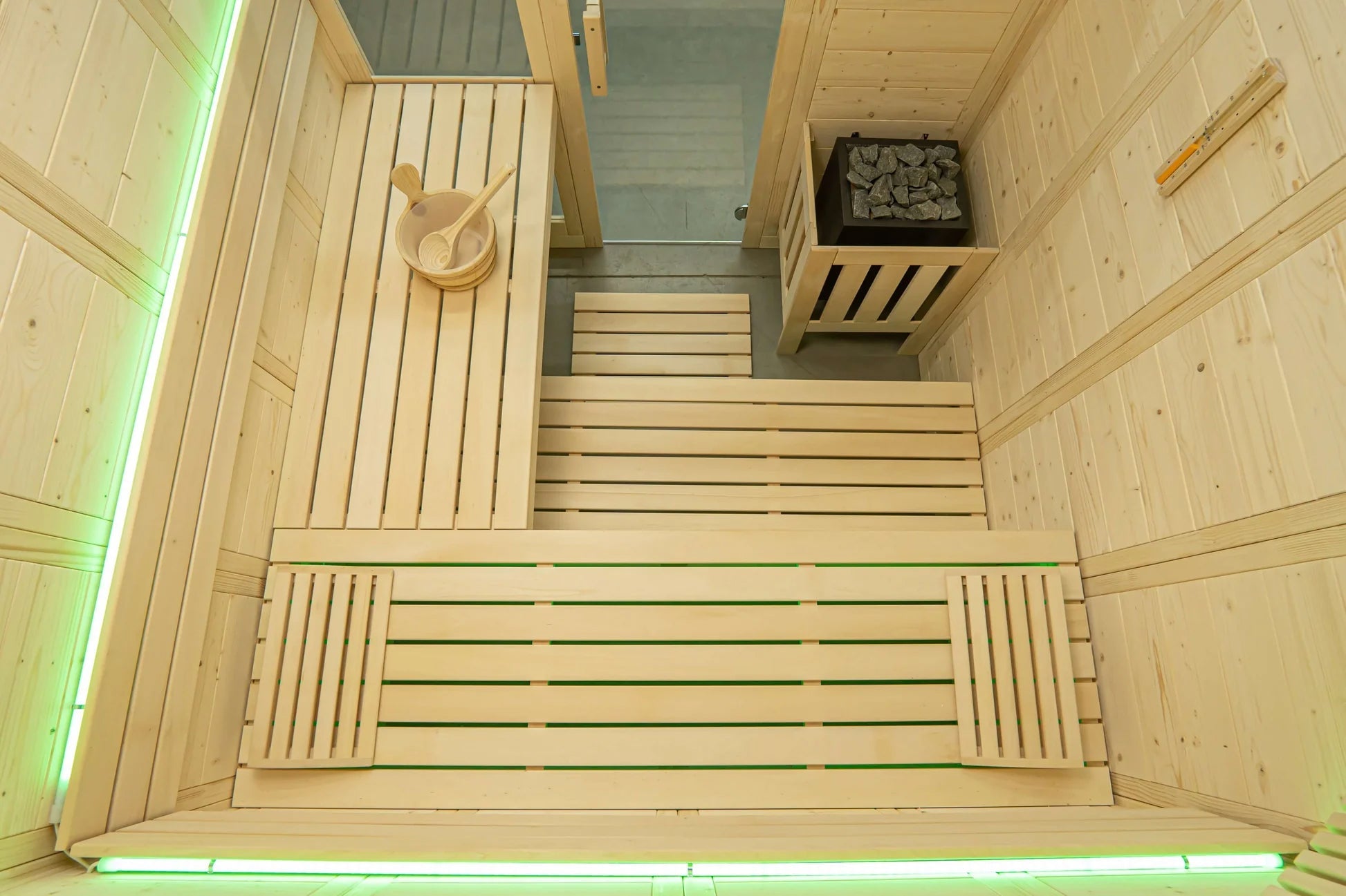 Interior view of wooden sauna with slatted benches, sauna bucket and ladle on upper shelf, sauna rocks in metal basket, and g
