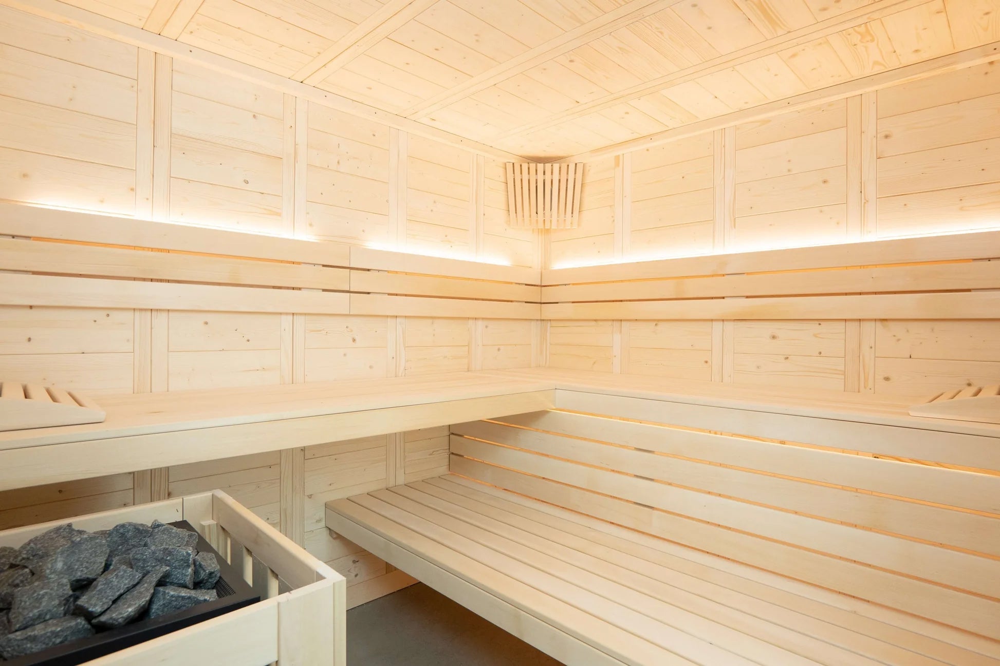 Interior view of light wood sauna with built-in benches, wooden paneled walls, LED lighting, and heated stone basin