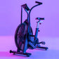 Blue and black air bike with large fan wheel, upright handlebars, and pedals on purple gradient background