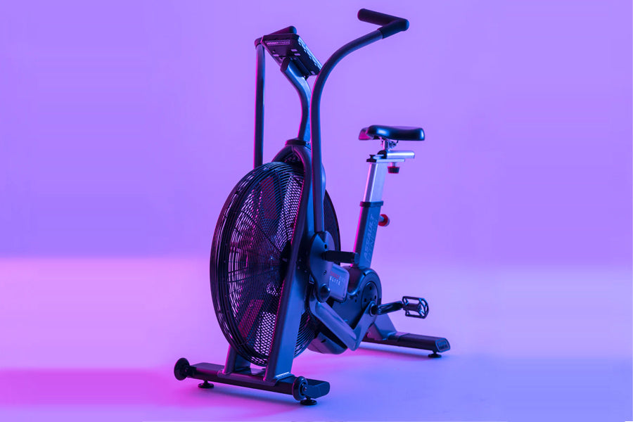 Blue and black air bike with large fan wheel, upright handlebars, and pedals on purple gradient background