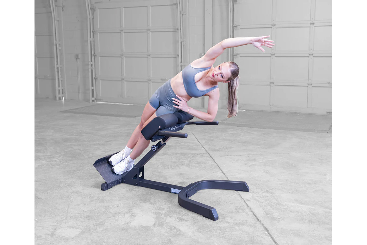 Woman performing side stretch on black Body-Solid 45-degree ab/back hyperextension machine, wearing blue sports bra and black