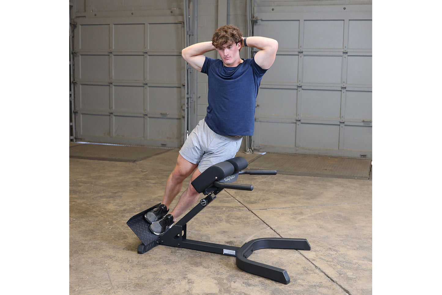 Man wearing navy t-shirt and gray shorts using black 45-degree ab/back hyperextension bench in garage setting