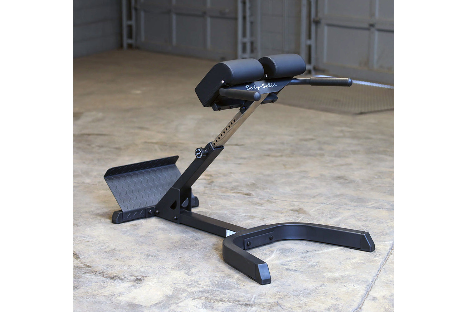 Body-Solid 45-degree ab and back hyperextension bench with black frame, padded backrest, and leg extension on concrete floor