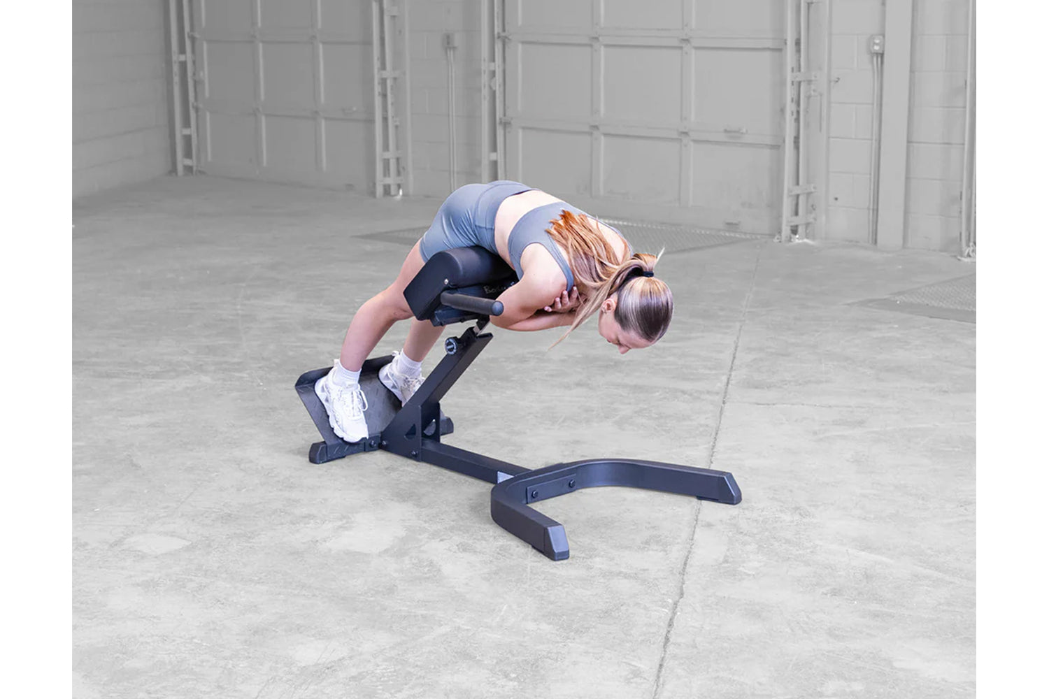 Woman in blue and white athletic wear performing ab exercise on black hyperextension bench in concrete garage setting
