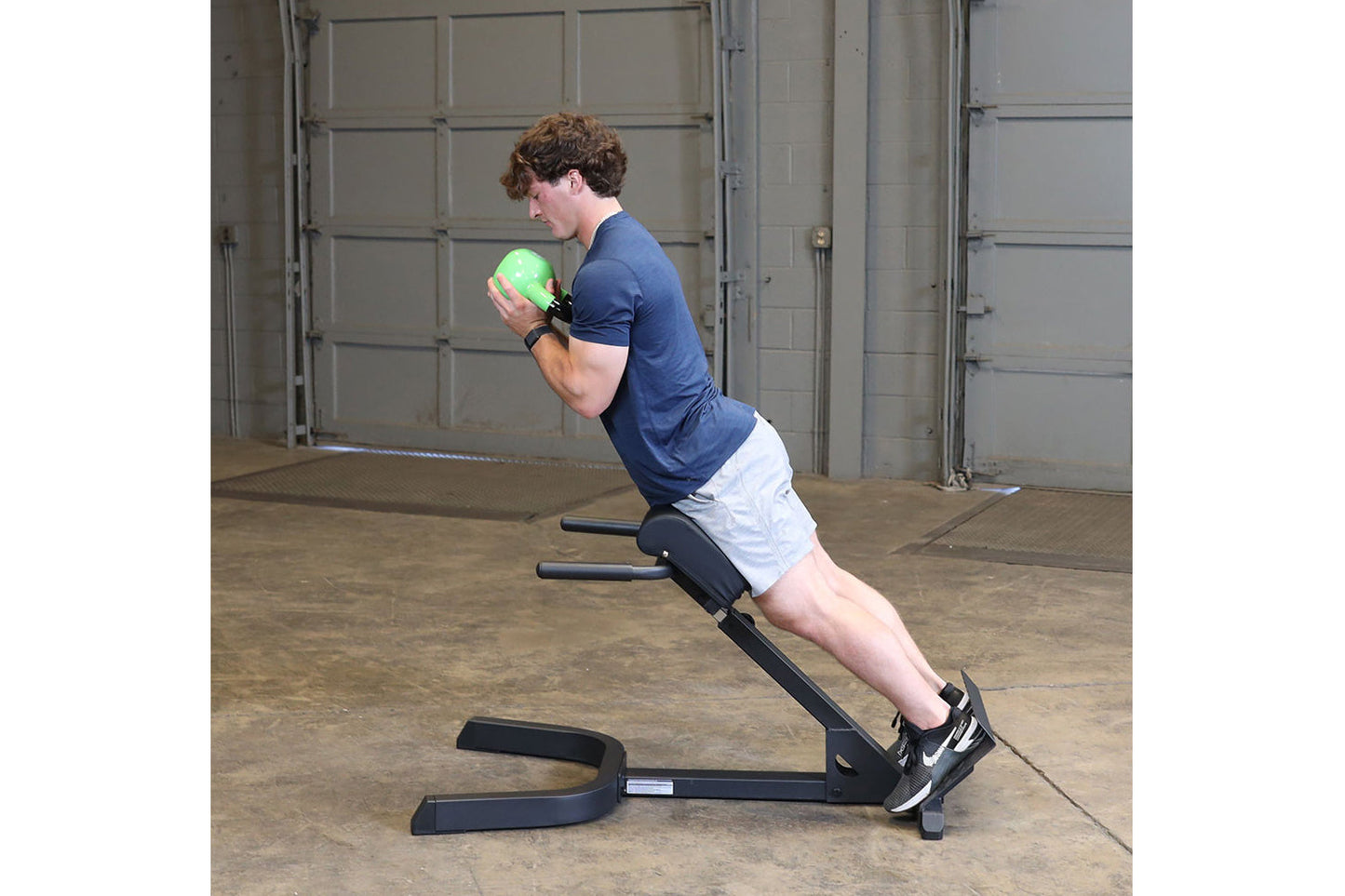 Man performing abdominal exercise on black Body-Solid hyperextension bench, holding green medicine ball, wearing navy t-shirt