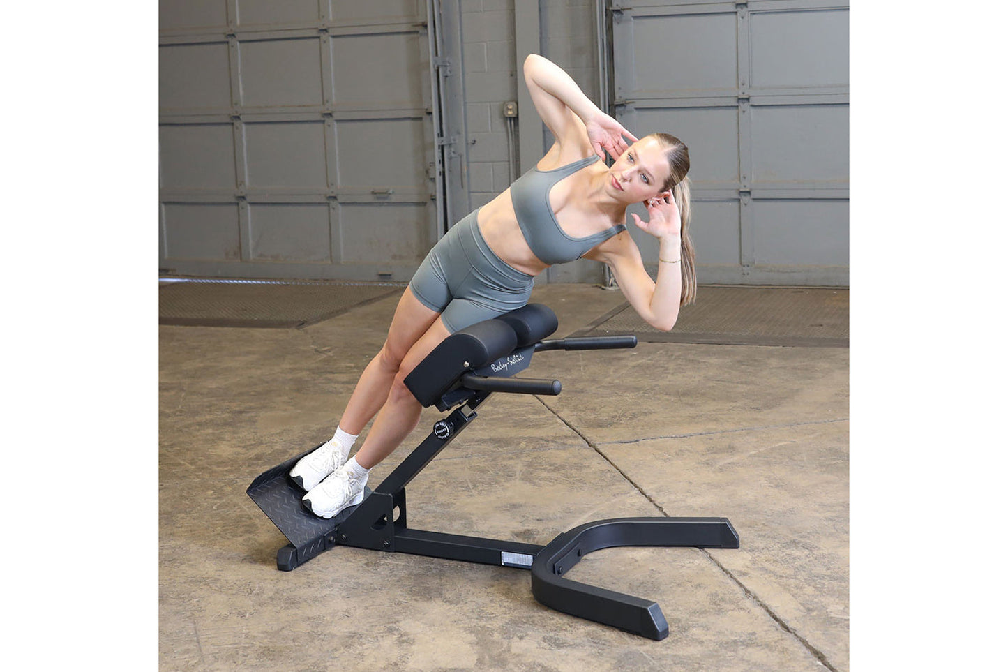 Woman performing ab crunch exercise on gray Body-Solid hyperextension bench in industrial gym setting