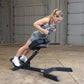 Woman performing abdominal exercise on black Body-Solid hyperextension bench in industrial gym setting, wearing gray sports b