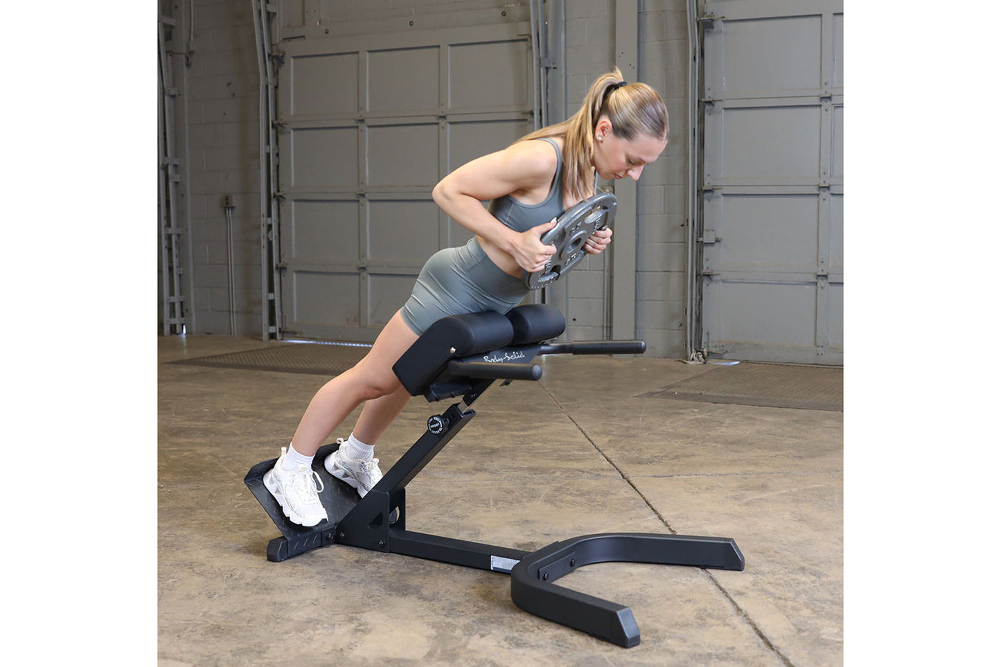 Woman performing abdominal exercise on black Body-Solid hyperextension bench in industrial gym setting, wearing gray sports b