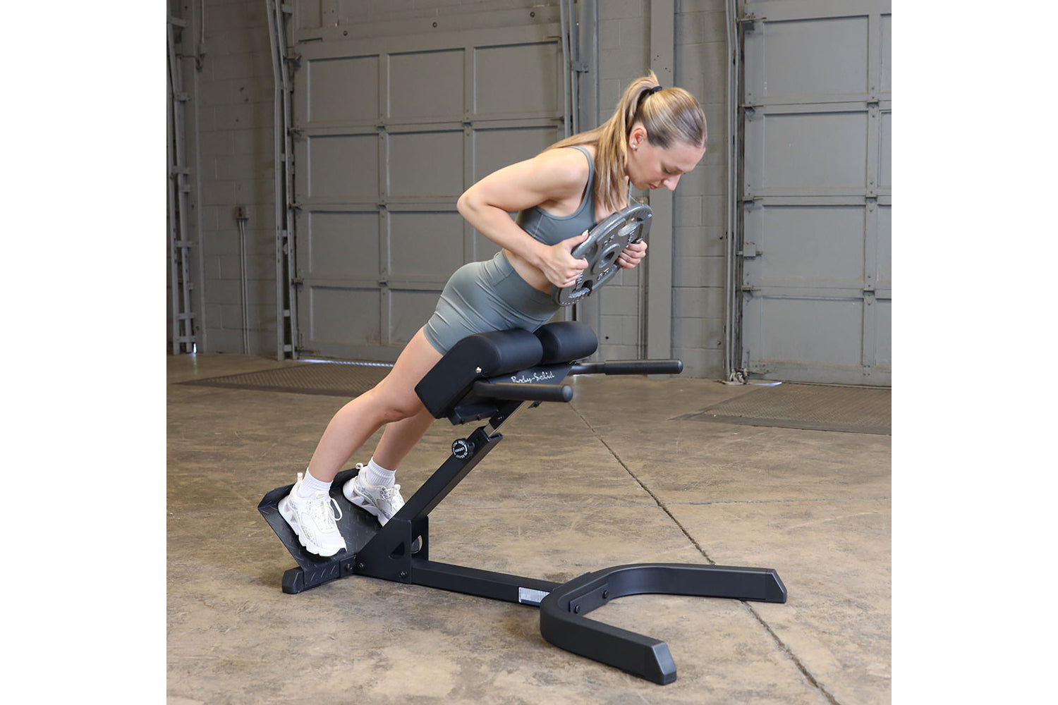 Woman performing abdominal exercise on black Body-Solid hyperextension bench in industrial gym setting, wearing gray sports b