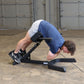 Person using black hyperextension machine with blue shirt and white tank top, performing abdominal exercise on concrete floor