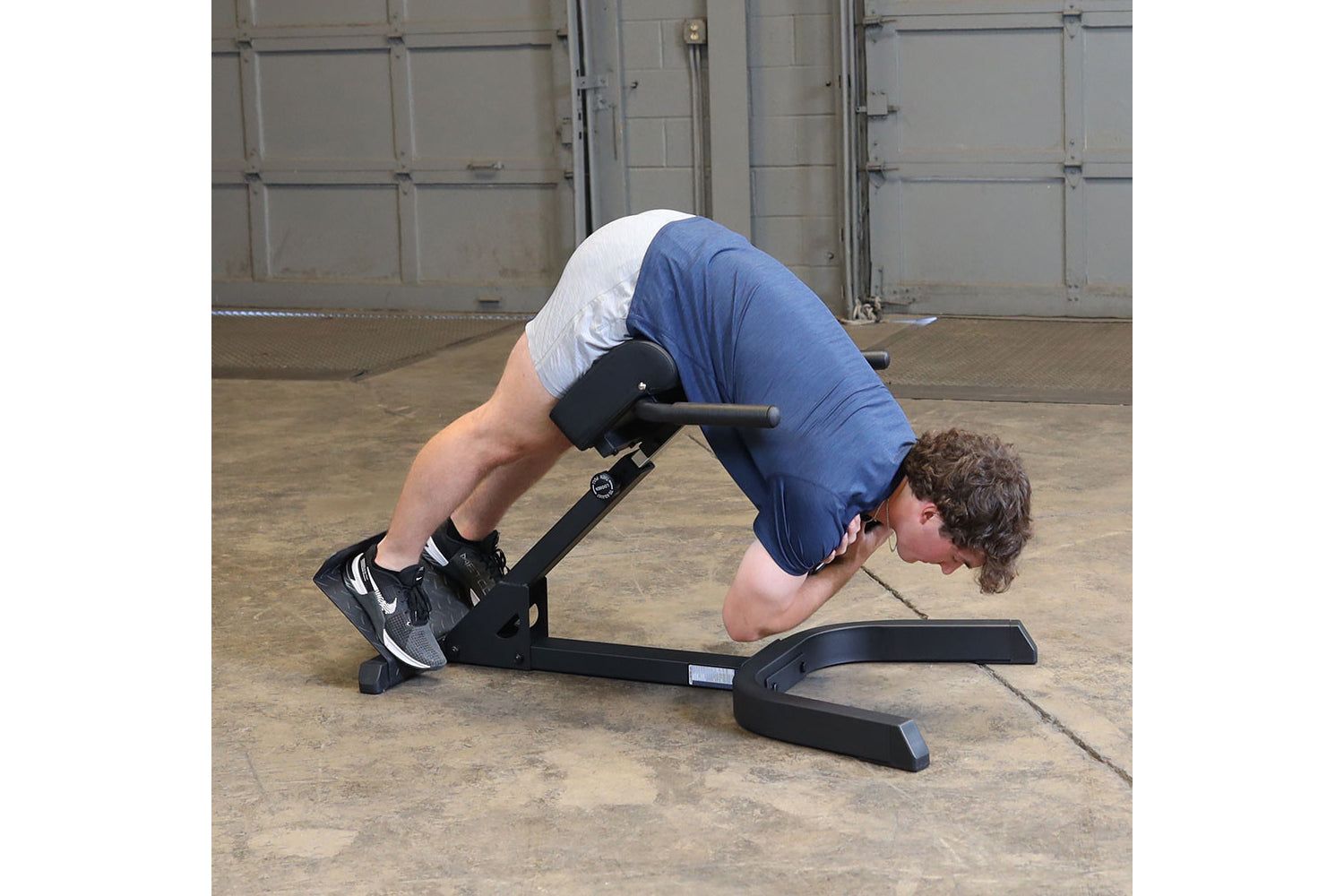 Person using black hyperextension machine with blue shirt and white tank top, performing abdominal exercise on concrete floor