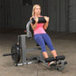 Woman using black and gray ab and back machine, wearing pink tank top and navy capri leggings, in gym setting
