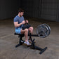 Man in blue t-shirt and light blue shorts using black seated calf raise machine with weighted plate in industrial gym setting