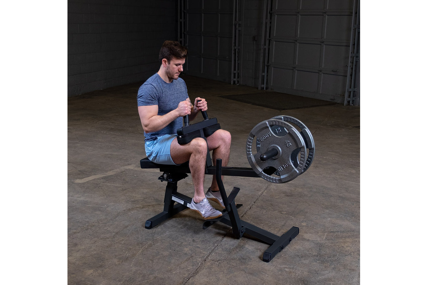 Man in blue t-shirt and light blue shorts using black seated calf raise machine with weighted plate in industrial gym setting