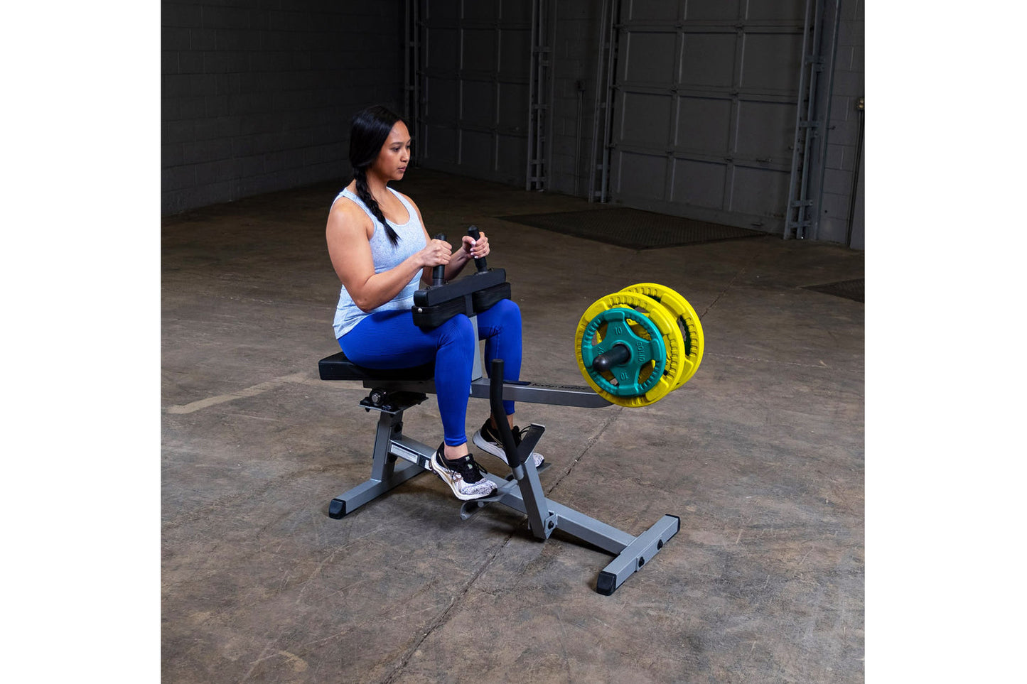 Woman using commercial seated calf raise machine with yellow weight plates in industrial gym setting
