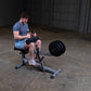 Man in blue t-shirt and light blue shorts using black and gray seated calf raise machine with weight plates in gym