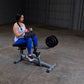 Woman performing seated calf raise on black and gray commercial gym machine with attached weight plate in industrial warehous