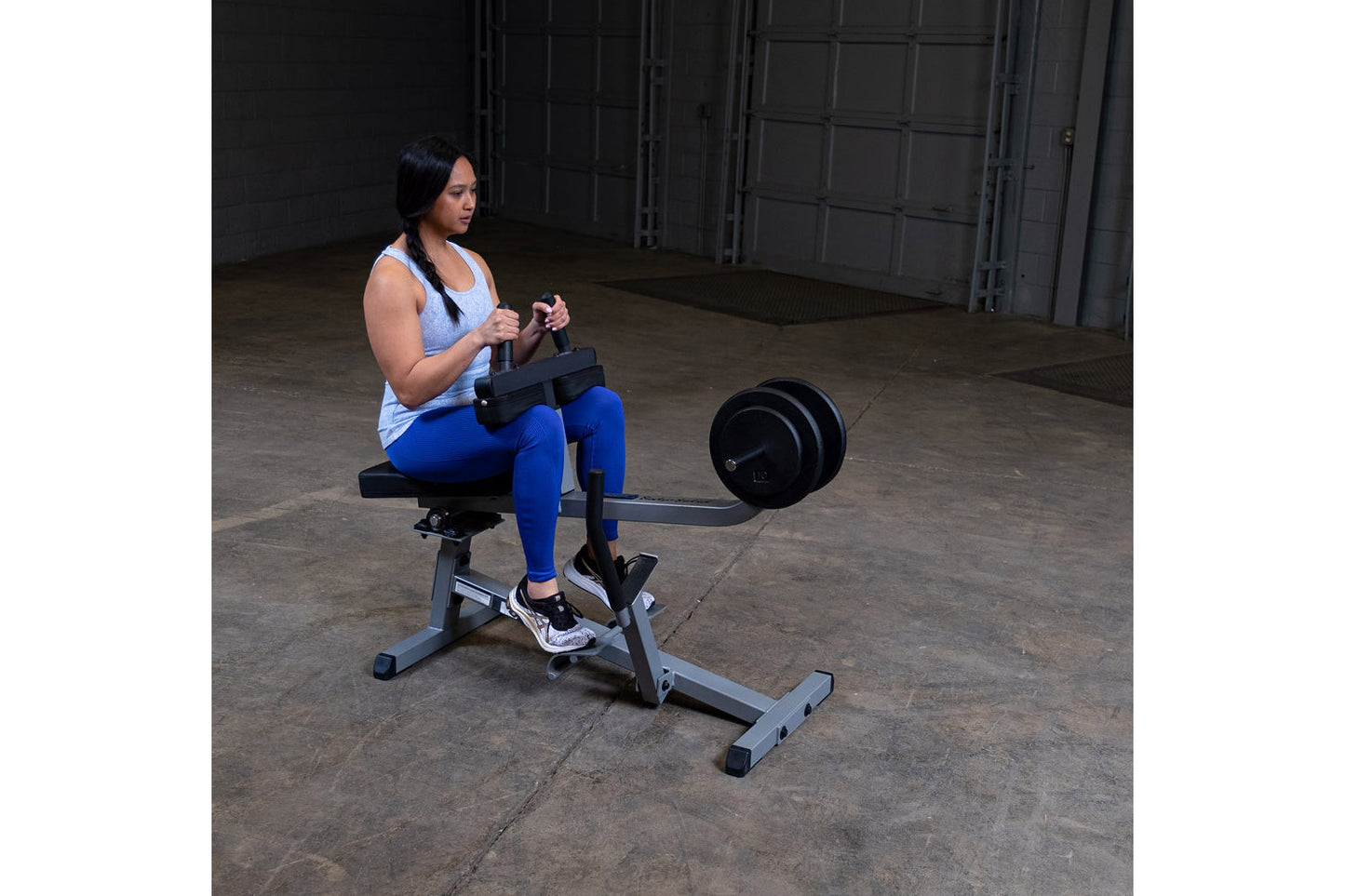 Woman performing seated calf raise on black and gray commercial gym machine with attached weight plate in industrial warehous