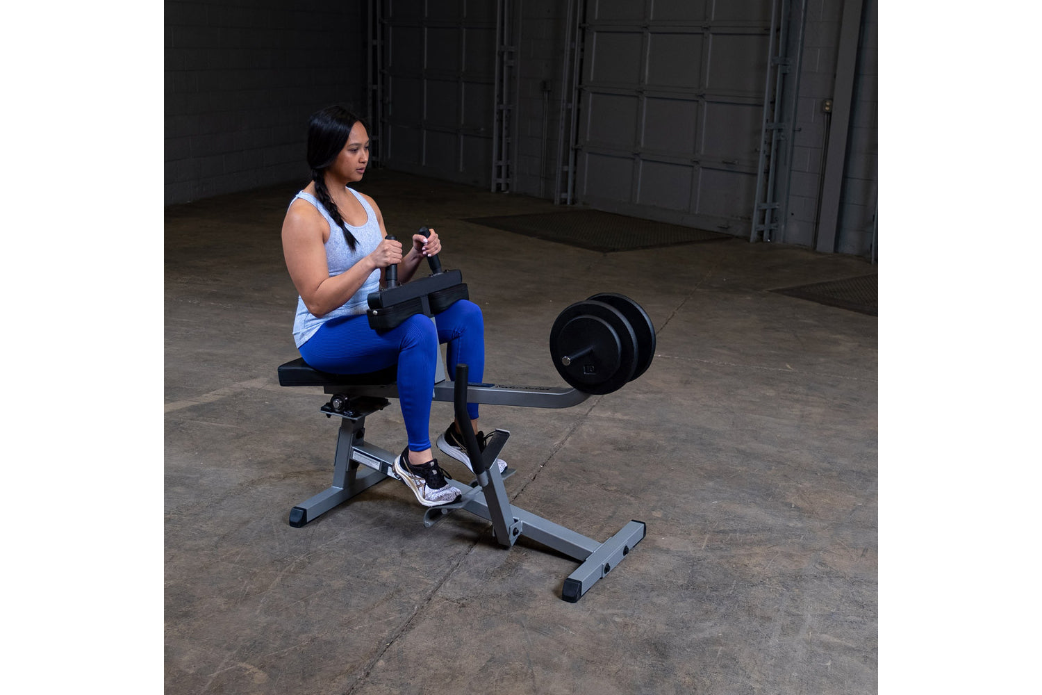 Woman performing seated calf raise on black and gray commercial gym machine with attached weight plate in industrial warehous