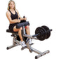 Woman using Body-Solid commercial seated calf raise machine with black weight plates, gray frame, and padded seat on white ba