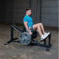 Man in blue t-shirt and gray shorts using black and gray leg press machine with loaded barbell plate in industrial gym settin