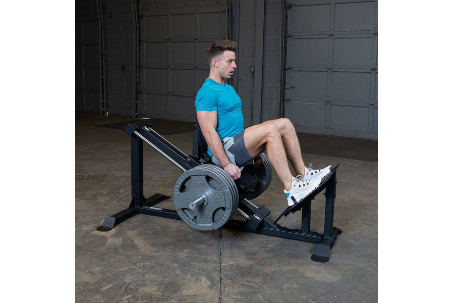 Man in blue t-shirt and gray shorts using black and gray leg press machine with loaded barbell plate in industrial gym settin