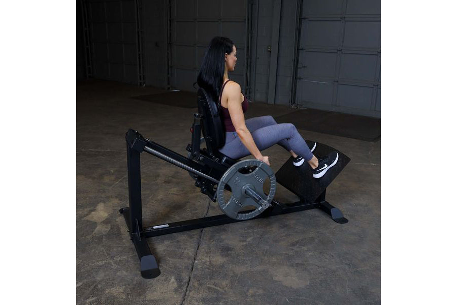Woman using black leg press machine with weighted plate, wearing purple leggings and black sneakers in gym setting